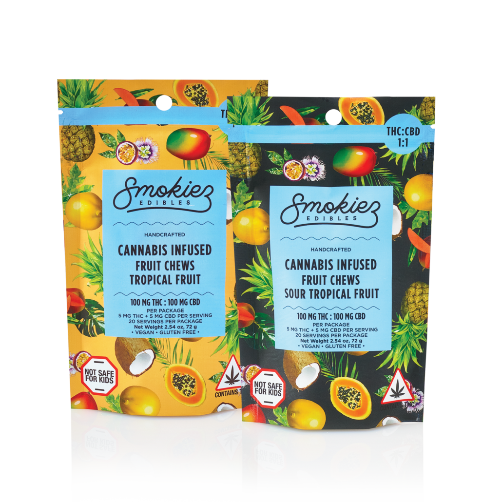 Two colorful packages of Smokiez Edibles cannabis-infused fruit chews are shown. The left package is bright yellow with tropical fruit illustrations and labeled "Tropical Fruit," while the right package is black with similar fruit illustrations and labeled "Sour Tropical Fruit." Both packages indicate 100 mg THC and 100 mg CBD content, with a warning symbol and "Not Safe for Kids" text. The design includes images of pineapple, mango, papaya, and coconut